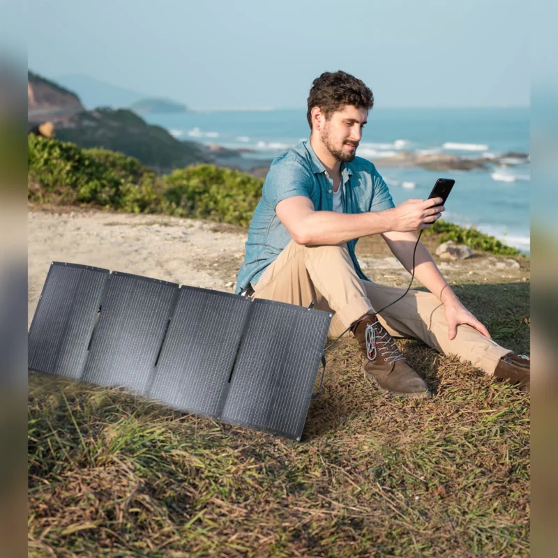 Hombre revisando su celular junto al panel solar Choetech SC006 desplegado sobre el pasto, frente al mar
